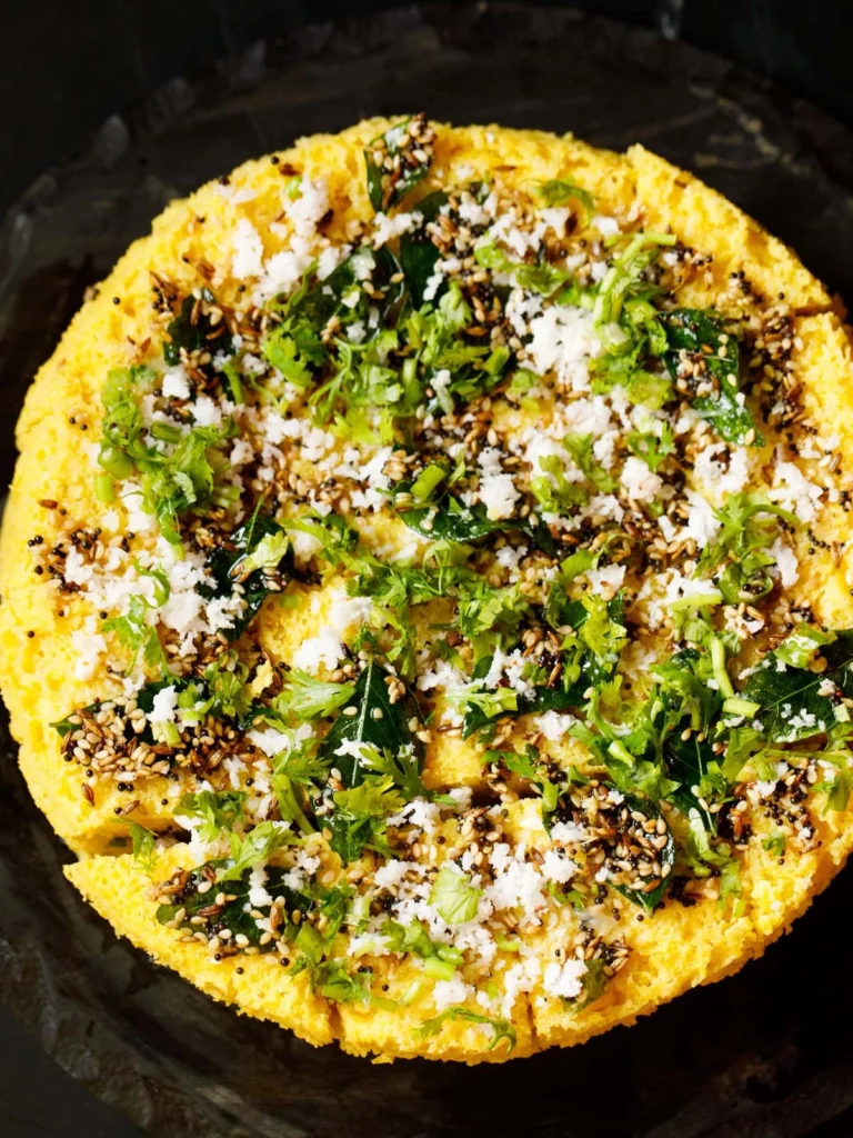 Soft and spongy squares of Gujarati dhokla garnished with mustard seeds, fresh coriander, and a light tempering, neatly arranged on a plate with chutneys in the background.