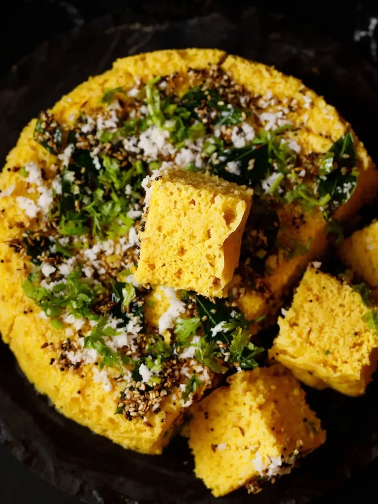 Close-up of fluffy yellow dhokla squares topped with mustard seeds, coriander, and sev, plated on a rustic dish.