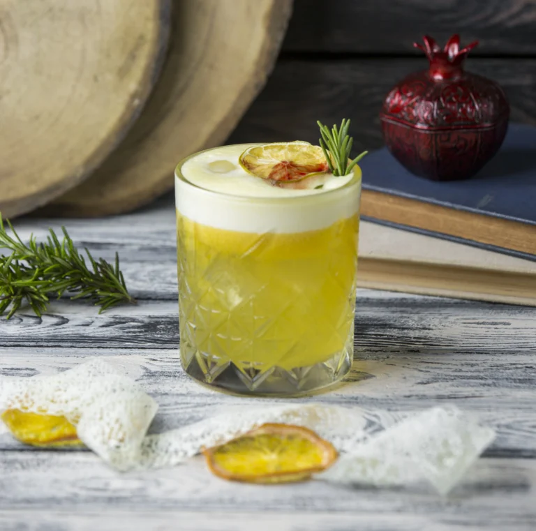 A refreshing yellow cocktail served in a crystal glass, garnished with a slice of dried lemon and a sprig of rosemary, placed on a rustic wooden table with dried fruit and books in the background.