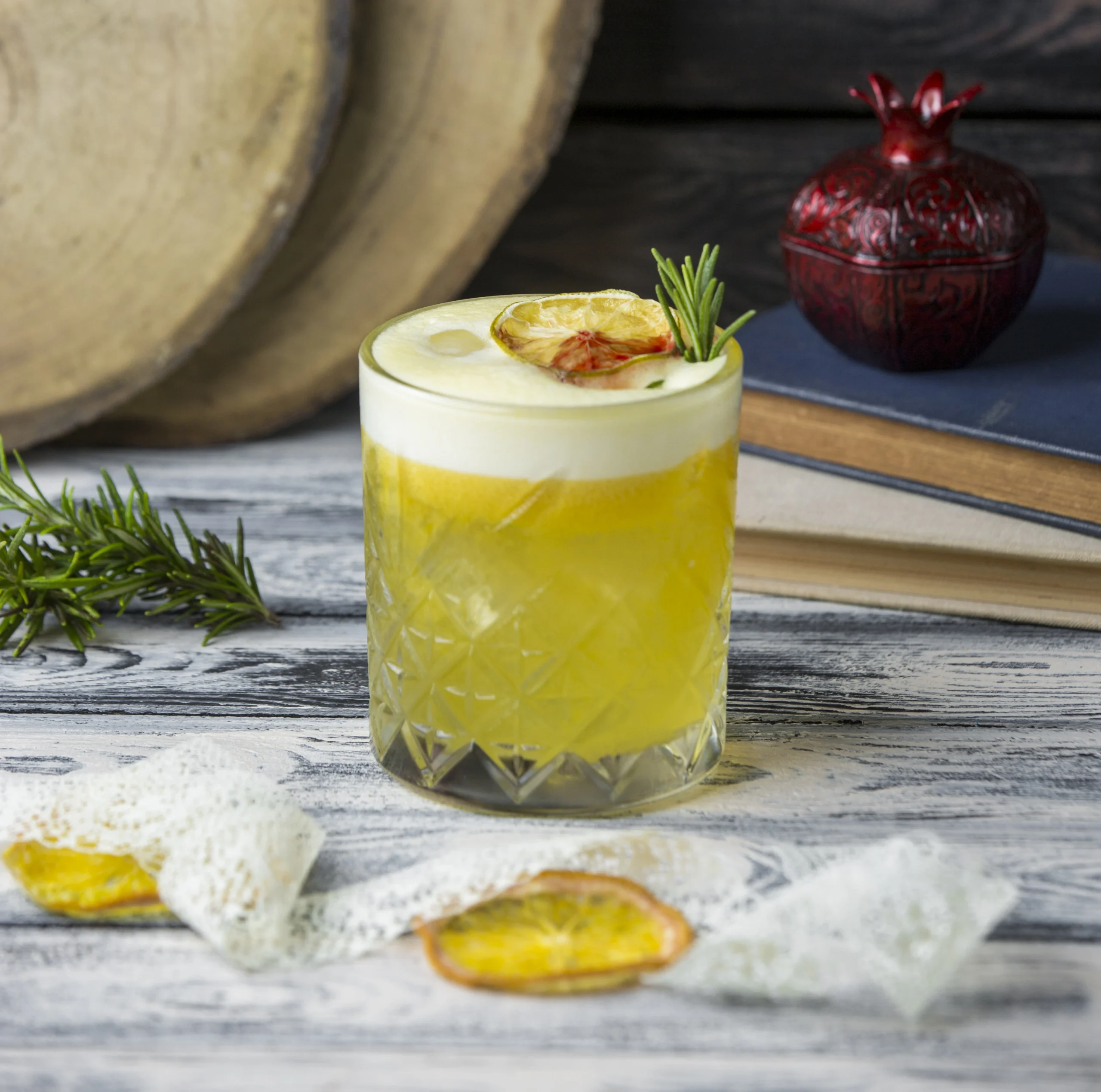 A refreshing yellow cocktail served in a crystal glass, garnished with a slice of dried lemon and a sprig of rosemary, placed on a rustic wooden table with dried fruit and books in the background.