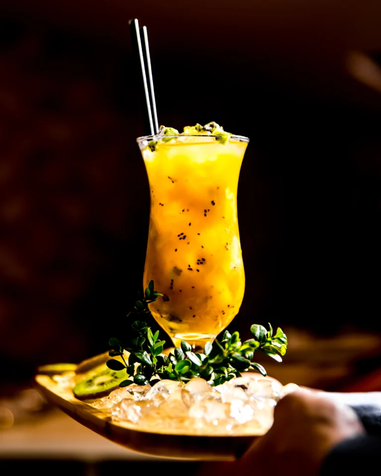 Spicy Mango Caipirinha cocktail served in a tall glass with black seeds, garnished with herbs and kiwi on crushed ice.