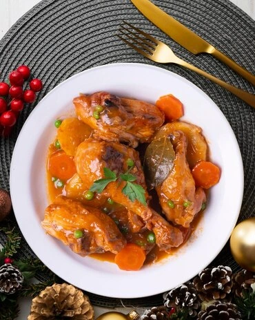 Chicken and apricot tagine served in a white bowl with carrots, peas, and a rich sauce, garnished with parsley and surrounded by festive holiday decorations.