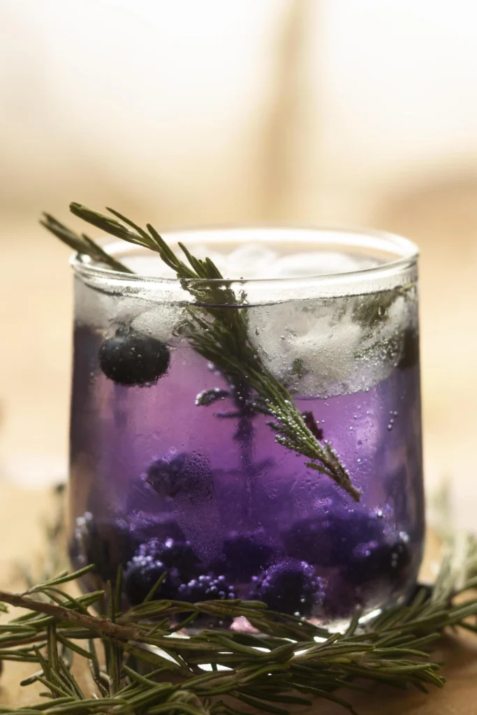 Purple spritz cocktail with butterfly pea flowers and lime on a white table.