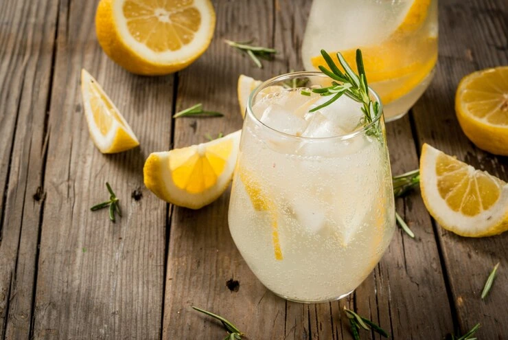 A bright yellow cocktail served in a textured crystal glass, topped with frothy foam, garnished with a slice of dried lemon and a sprig of rosemary, set on a rustic wooden table with books and herbs in the background.