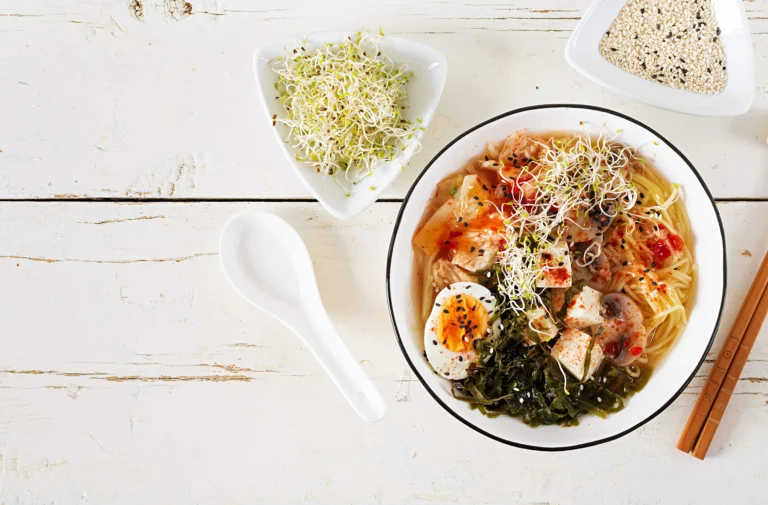 Miso ramen with kimchi, seaweed, egg, tofu, mushrooms, and sprouts in a white bowl on a rustic white wooden table.