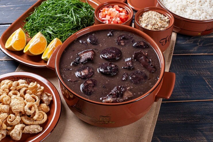 Homemade Brazilian Feijoada in a clay pot with sides of rice and orange slices.
