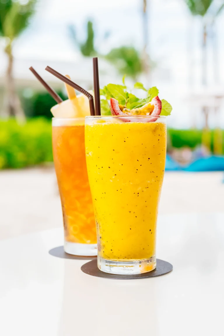 A tall glass of chilled mango lassi garnished with mint and fruit, served with a straw on a sunny outdoor table.