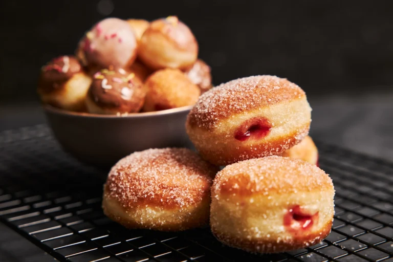 Close-up of sugar-coated Bolinho de Chuva filled with jam, with a bowl of assorted doughnut balls in the background.