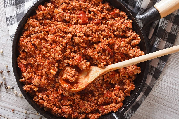 A close-up of a bowl of pasta with thick, rich ragu sauce, featuring visible ground meat and tomato chunks, with a fork twirling spaghetti above the bowl.