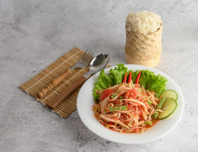 Thai green papaya salad served on a white plate with fresh vegetables and lime slices, accompanied by sticky rice in a bamboo container.