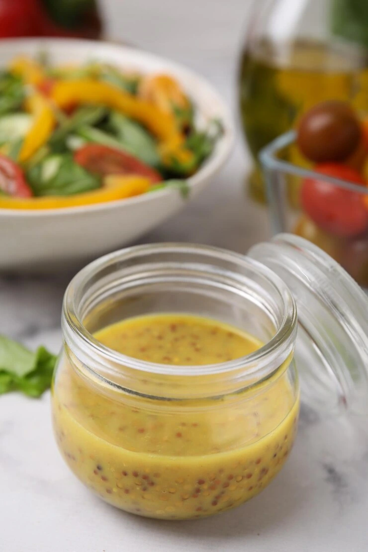 A glass jar filled with lemon vinaigrette surrounded by fresh ingredients including tomatoes, parsley, oil, vinegar, and a whisk on a white wooden table.