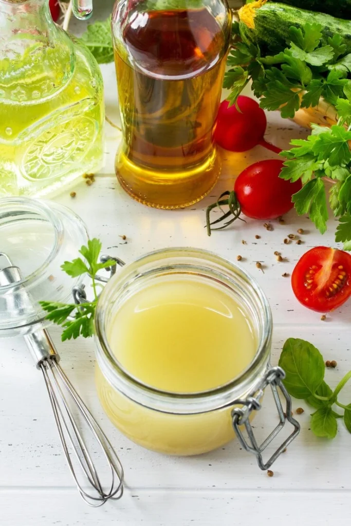A glass jar filled with homemade lemon vinaigrette placed on a white wooden table, surrounded by fresh cherry tomatoes, parsley, basil leaves, a whisk, and bottles of olive oil and vinegar.