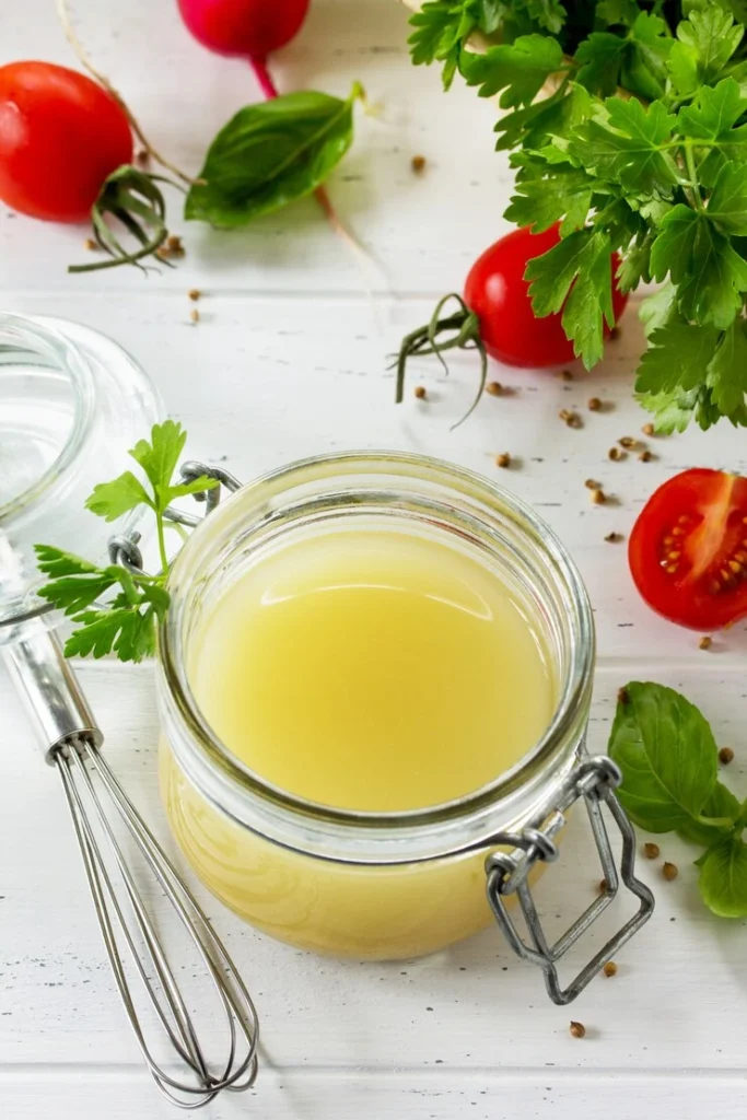 A glass jar filled with homemade lemon vinaigrette placed on a white wooden table, surrounded by fresh cherry tomatoes, parsley, basil leaves, a whisk, and bottles of olive oil and vinegar.