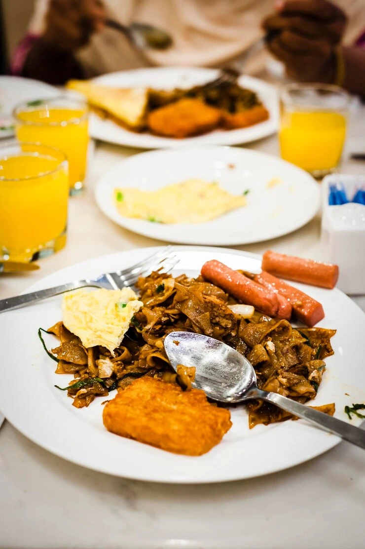 Close-up of a Almoço de Domingo plate with stir-fried noodles, omelette, sausages, and a hash brown, served with glasses of orange juice on a white table.