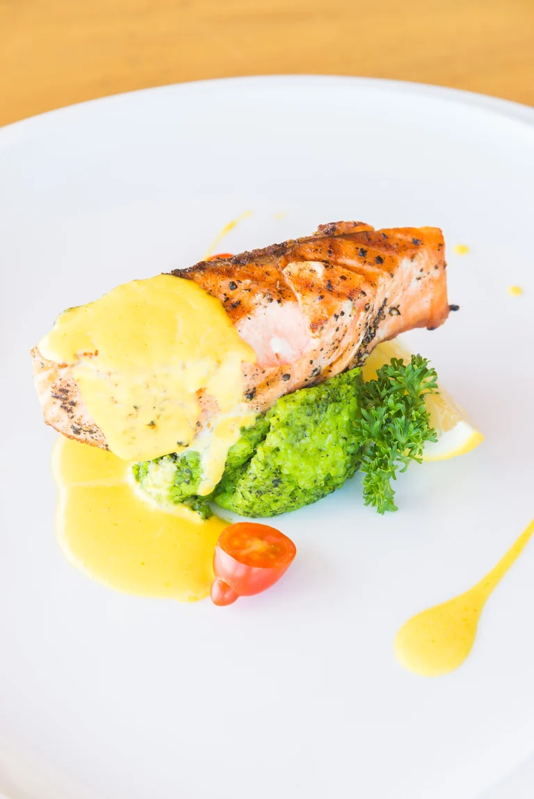 Grilled Wild Alaskan Salmon steak with creamy yellow sauce, green vegetable mash, and tomato garnish on a white plate.