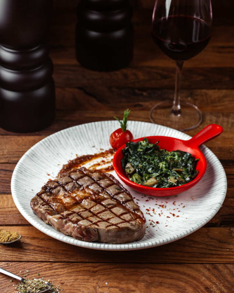 Juicy Grilled Ribeye Steak served with sautéed greens and a glass of red wine on a rustic wooden table.