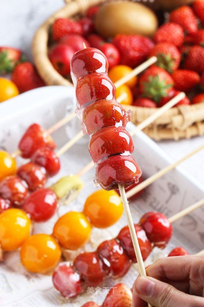 Close-up of homemade Tanghulu featuring glossy candied strawberry skewers with a hard sugar coating, held above a tray of colorful fresh fruit and additional candied fruit skewers.