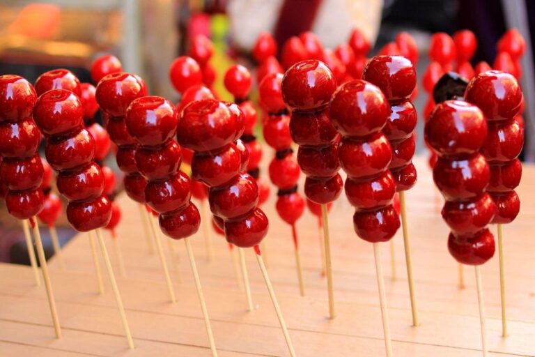 Glossy red tanghulu skewers made with candied fruit, likely cherries or hawthorn berries, stacked on wooden sticks and displayed upright on a wooden table with a softly blurred market background.